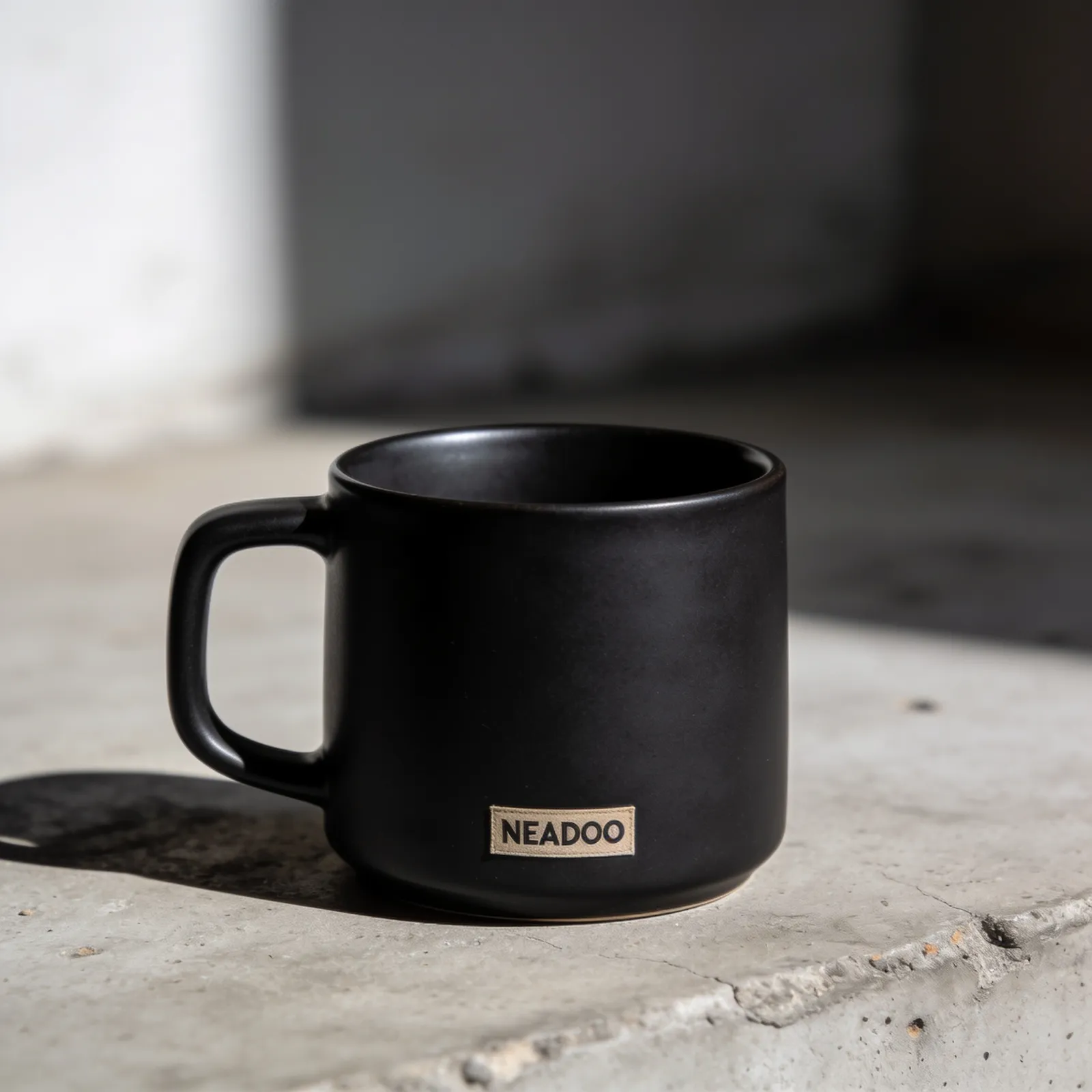 Czarny matowy kubek ceramiczny wygenerowany przez Seedream 4.5: beżowa naszywka materiałowa z napisem NEADOO, ostre światło słoneczne, betonowy parapet